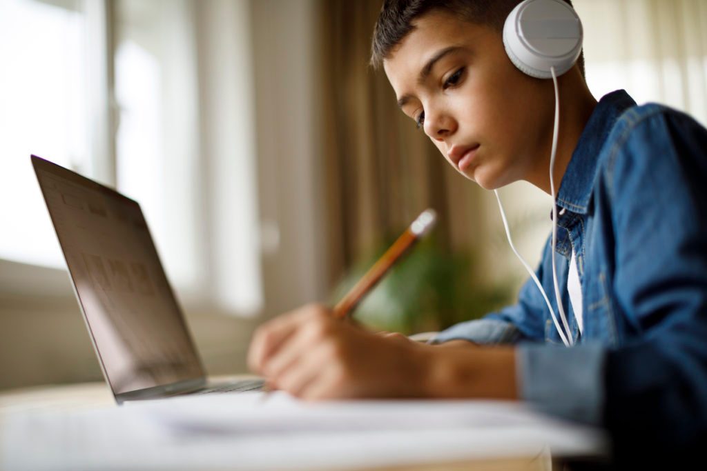 Youth with headphones writing in notebook in front of a laptop