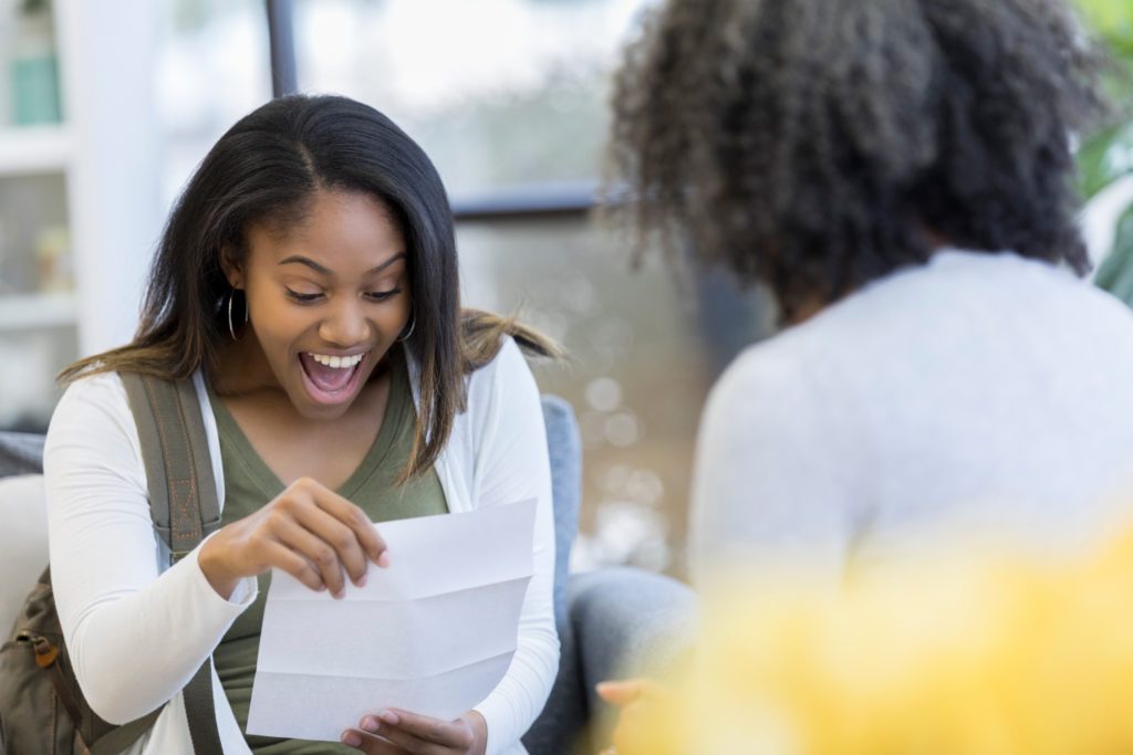 African American teenage girl is surprised and delighted to receive a college acceptance letter. Her guidance counselor is sitting in the foreground.